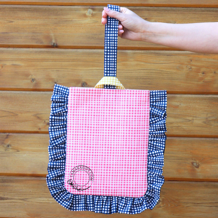 A hand holding a book bag with a pink and blue checkered pattern and yellow ruffles, against a wooden background.