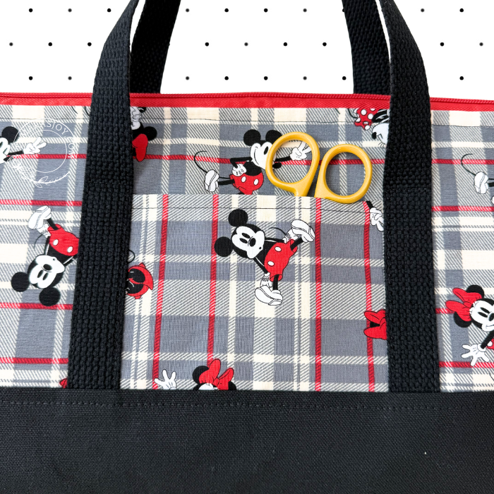 A close-up of a tote bag featuring a gray, white, and red plaid pattern with scattered Mickey Mouse illustrations, black handles, and a black bottom panel. A pair of yellow-handled scissors rests on the front pocket.