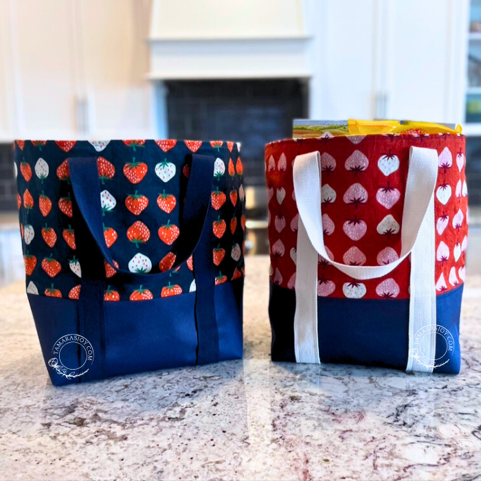 Two heavy duty grocery tote bags with a geometric strawberry pattern, one in blue with red hearts and the other in red with blue hearts, displayed on a countertop.