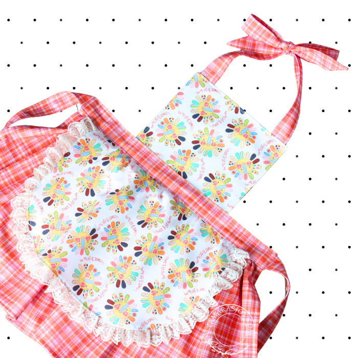 A child's apron with a floral pattern and red checkered trim, featuring a bow tie at the neck.